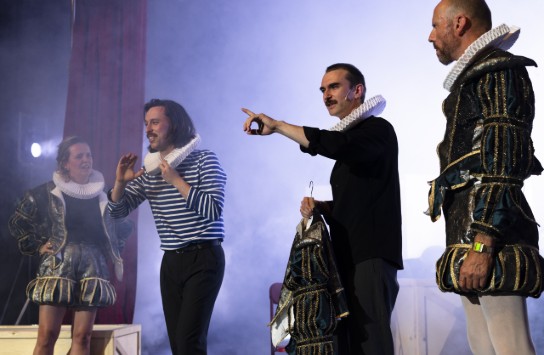 Vier acteurs op toneel, twee in historische kostuums met kraag, één in gestreept shirt, één wijzend met hand. (vergroot foto)