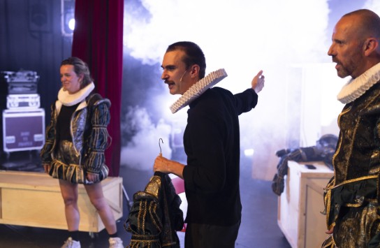 Drie acteurs in renaissancekostuums met ruchekragen op een theaterpodium met rook en licht. (vergroot foto)