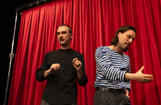 Twee mannen met microfoons op het podium voor een rode theatergordijn, één in zwart, de ander in blauw-wit gestreept. (vergroot foto)