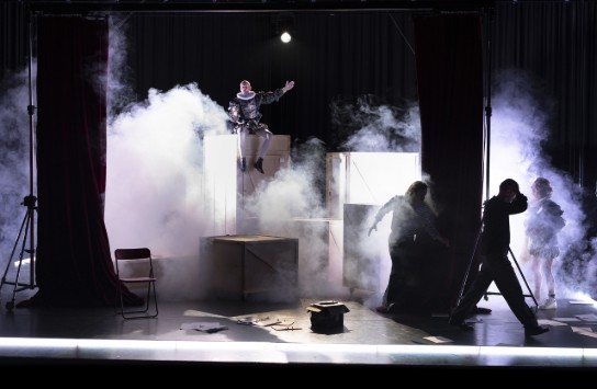 Theaterpodium met rook, een acteur in renaissancekostuum op kisten, en drie andere personen in schaduw achter gordijnen. (vergroot foto)