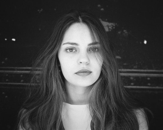 Zwart-wit portret van een jonge vrouw met lang haar, neutrale gezichtsuitdrukking en een donkere achtergrond. (vergroot foto)