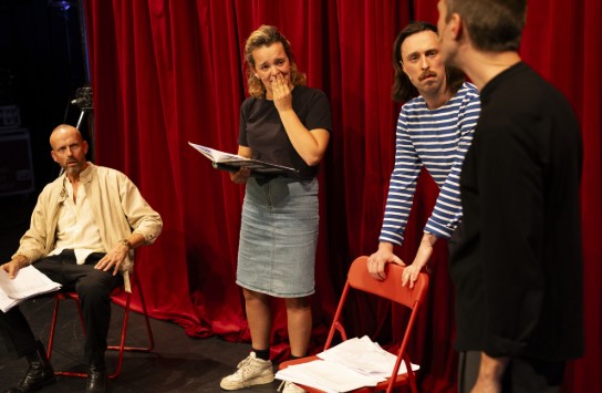 Vier acteurs oefenen een scène met rode gordijnen op de achtergrond, één vrouw leest uit een script. (vergroot foto)