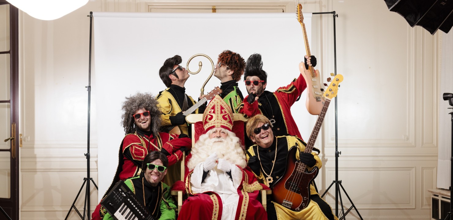 Sinterklaas zit op een stoel, omringd door zes mannen in kleurrijke kostuums met muziekinstrumenten.