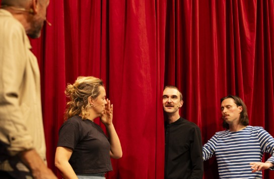 Vier acteurs oefenen een scène achter een rood theatergordijn met scripts en stoelen op het podium. (vergroot foto)