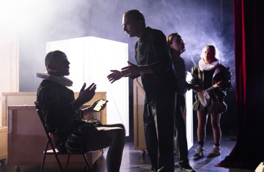 Vier acteurs in een donkere theateromgeving, twee in historische kostuums, één met telefoon, in gesprek achter het podium. (vergroot foto)